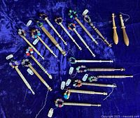 Full lot of 18 varied wooden lacemaking bobbins arranged on blue velvet, most with colorful glass bead spangles acting as weights.