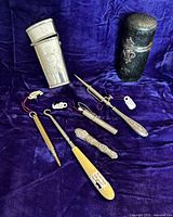 Overview of complete lot showing leather companion case, assorted tools including needlecases, button hook, punch, and chatelaine pencil on purple velvet background.