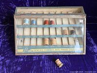 Front view of the Lily metal thread display case showing original spools in divided compartments with colorful thread spools and printed color labels inside transparent hinged glass lid.