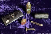 Top-down view of six items including WWII black leather sewing kit case, WWI leatherette kit case, small steel flask kit, army-green metal canister, folding cutting tool and metal clip.