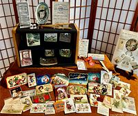 Full view of the lot showing a large assortment of colorful vintage needlecases, needlebooks, advertising cards, novelty items spread out in front of a black and wood-framed display vitrine.