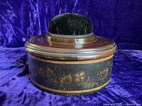 Side view of round wooden box decorated with bronze powder stencil floral motif and painted bands, topped with a black velvet pincushion lid.