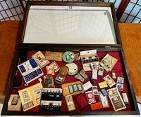 Wide view of dark wooden hinged glass vitrine containing multiple needlecases and sewing needle packets arranged on red fabric.