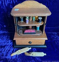 Wooden thread stand with tambour doors open revealing swing-out thread holders with multiple thread spools and pincushion on bottom shelf. Drawer below holds sewing supplies. Two decorative ear-of-corn pinholders in front.