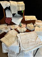 Wide shot of assorted linen textiles stacked and draped over furniture including napkins, tablecloths, and towels in white and cream shades with visible embroidery and damask patterns.