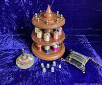 Full view of two-tier wooden thread stand with thread spools, German pinkeep, plaid velvet pincushion, and assorted antique thimbles arranged on deep blue velvet background.