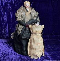 Two antique half-doll pincushions together against purple velvet background, showing difference in size and color of attire.