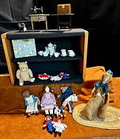 Wide view showing all items in lot: two Singer sewing machine toy banks, multiple stuffed dolls including Raggedy Ann and Andy, small child's tea set in box, knitted teddy bear, and braided doll house rug.
