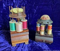 Two antique wooden thread holders on blue fabric background. Larger holder has green, pink, red thread spools on spindles and a yellow patterned pincushion on top. Smaller has multiple small spools and a multicolor brocade pincushion.