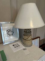 Art print with silver frame next to the lamp with a white shade and woven wicker base on a table.