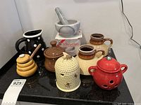 Photo showing ceramic bee hive honey pots, creamer jug, three mugs, marble mortar and pestle on a black surface