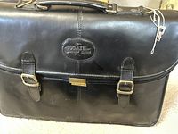 Front view of black leather briefcase with Bugatti logo embossed on an oval patch, two front buckled straps, and gold-tone central lock.