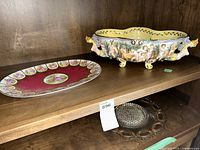 Shelf display showing the Capodimonte porcelain bowl with cherubs and a vintage German glass plate with detailed painted scenes