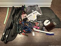 Photo showing a collection of automotive items including booster cables, car mats, brushes, ice scrapers, paperwork, and cleaning accessories on the floor.