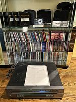 Photo of Sherwood CDC-690T CD player on floor with manual on top, mirrored shelf with large CD collection behind and some electronics on shelf above.