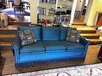 Front view of the teal blue-green Lori Morris sofa with two patterned cushions and fringed base.