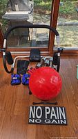 Ab Rocker device with seat damage, red exercise ball, ankle and wrist weights, hand weights, and motivational sign arranged on wooden floor by window.