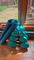 View of the blue yoga mat rolled and placed next to a black dumbbell stand with teal plastic dumbbells on a hardwood floor near a window.