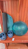 Photo shows teal Lululemon yoga mat rolled up with green exercise ball and two blue hand weights on a wooden bench.