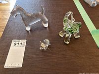 Three glass animal sculptures: two elephants with green internal highlights and one clear textured Scottie dog on a leather surface with a maxsold lot tag.