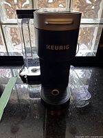 Front view of the black Keurig K-Slim coffee maker showing the water reservoir and a transparent accessory.