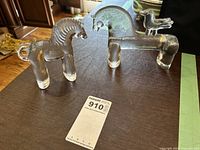 Two clear glass horse sculptures on a dark surface; one smaller horse and one larger horse with a bird on its back