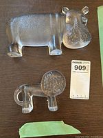 Top down photo showing both the hippo and lion glass sculptures placed on a dark wood surface with a MaxSold tag nearby.