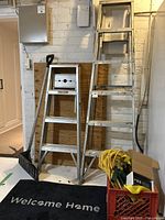 Two metal step ladders, a snow shovel, red crate with cords and hardware, and black welcome mat shown against a white brick wall.