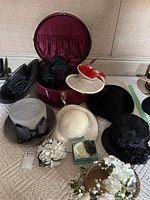 Photo of eight ladies hats and fascinators in black, grey, cream and white colors, with various flower and feather decorations, plus a maroon round travel hat box.