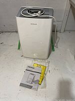 Front view of the white Noma dehumidifier with power cord placed on a cement floor next to the manual in plastic packaging.