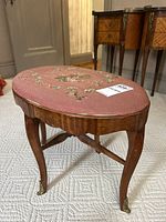 Front side view of oval needlepoint stool showing pink floral embroidered top, veneer wood base, and curved legs with brass feet.