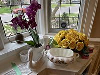 Photo showing ceramic swan planter, orchid planter with artificial orchid, yellow artificial roses, white ceramic mug, tin pitcher, and plant food container arranged on a window sill.