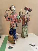 Two porcelain Asian female figures in traditional colorful attire, one standing on a brown decorative base, approximately 12 inches tall.