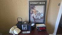 Full lot including poster, two clock radios, dial phone, and black item (CD player with speakers and remote) on table.