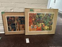 Two framed modern art prints leaning against a white brick wall on the floor, left print abstract urban scene by Prevost, right print colorful abstract figures by Zarfati