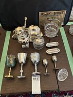 Silver plated items arranged on a table including goblets, cream and sugar set, candy dish with glass insert, glass dresser jar with silver plate lid, shaker, carving utensil rests, and more.