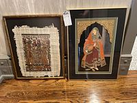 Two framed artworks side by side; left is a mixed media with papyrus background and gem paint, right is a framed papyrus painting of a woman in traditional attire with gold decoration.