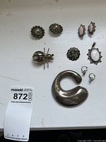 Top-down photo showing entire lot of sterling silver jewelry arranged on a white surface including bug pin, clip-on earrings, pink stone set pendant and earrings, black filigree brooch, and silver hoop earrings.