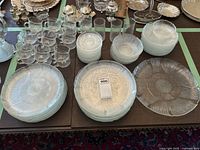 Overview of multiple glassware and dinnerware items arranged on table showing plates, bowls, glasses, mugs, and platters with floral pressed glass design.