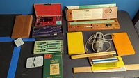 Photo showing an assortment of vintage architect drafting supplies including tool sets, rulers, books, and an electric eraser on a dark surface with blue tape markings.