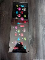 Full boxed set of 24 assorted vibrant colored blown glass Christmas ornaments by Robert Stanley in original packaging on wood floor.