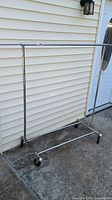 Full view of the metal clothing rack with four wheels on a concrete surface outside a beige siding wall.