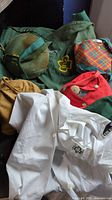 Photo showing multiple Girl Scout items including the green and red uniform jackets, white uniform blouse, canteens with colorful covers, and a green knapsack all with Girl Scout logos and patches visible.