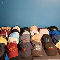 Photo showing 25 assorted hats arranged in rows on a black surface with various logos including places and advertising.