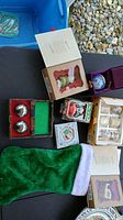 Photo showing multiple boxed Christmas ornaments including a green Christmas tree ornament, ball ornaments, and a green Christmas stocking on a table surface