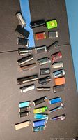 Top view of 28 assorted lighters arranged on a black surface, showing mixed shapes, colors and styles including refillable and disposable lighters.