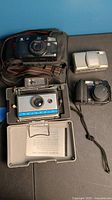 Lot of film and digital cameras on a blue and black background showing four cameras with the Polaroid camera case open