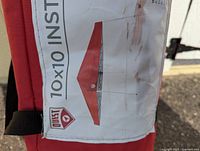 Red carrying bag for pop-up canopy with printed label showing 'QUEST 10x10 Instant Pop-Up Canopy Tent' and illustration of tent in red.