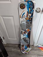 Full snowboard leaning against a door showing the attached grey bindings and colorful graphic design, condition visible with minor surface marks.