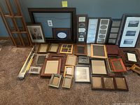 Photo showing about 25 different picture frames of varying sizes and colors arranged on carpeted floor against wall, including multi-photo collage frames and standalone frames.